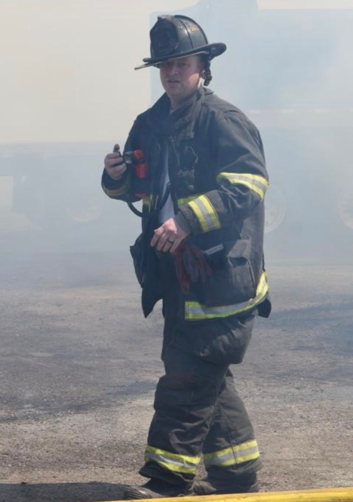 Bryan Syrett in full firefighter turnout gear and helmet walking through smoke outdoors, with a fire truck behind him.